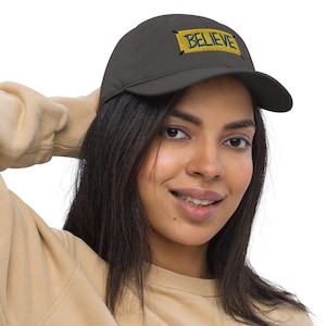 以下が含まれることがあります： 黄色と青で「BELIEVE」の文字が刺繍された濃いグレーの野球帽。帽子のデザインは前面の中央に配置されています。帽子は人がかぶっています。