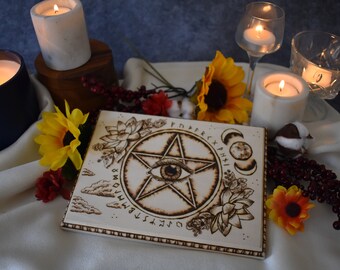 May include: A white wooden square tile with a wood-burned design of a pentagram with an eye in the center. The pentagram is surrounded by a circle with runes and symbols. The tile is surrounded by candles, sunflowers, and other decorative items.