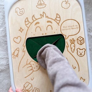 Puede incluir: Un juguete de madera con un diseño de monstruo de dibujos animados y un bolsillo de fieltro verde. El tablero tiene un marco blanco e incluye ilustraciones grabadas de un barco, una manzana, un pato, un corazón, un cohete, una zanahoria, un dado y un oso. El texto en un bocadillo dice "Was kitzelt in meinem Bauch?"