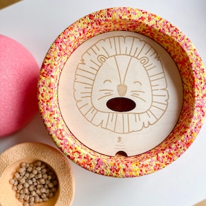 Può includere: Una ciotola di legno a forma di leone con un esterno macchiato di rosso, giallo e rosa. La ciotola è piena di piccole palline marroni. La ciotola è su una superficie bianca.