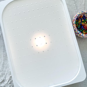 Può includere: Un vassoio di plastica bianco con una superficie perforata. Il vassoio ha angoli arrotondati e un bordo leggermente rialzato. Un piccolo contenitore colorato pieno di piccoli oggetti colorati è sullo sfondo.