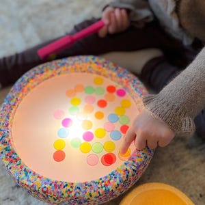 May include: A brightly lit children's toy with a speckled rim and a light-up surface. The surface displays an array of colourful, translucent circles. A child's finger points at the circles. A pink torch and a yellow bowl are also visible.