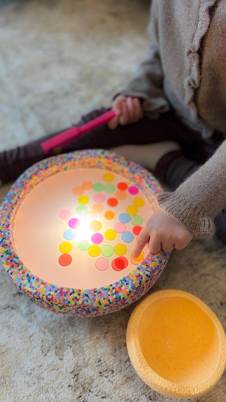 May include: A brightly lit children's toy with a speckled rim and a light-up surface. The surface displays an array of colourful, translucent circles. A child's finger points at the circles. A pink torch and a yellow bowl are also visible.