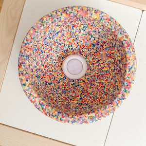 Könnte beinhalten: Ein gesprenkeltes Waschbecken mit einem hellfarbigen, kreisförmigen Ablaufdeckel. Das Becken weist ein Muster aus kleinen Punkten in verschiedenen Farben auf, darunter Rot, Gelb, Blau und Grün. Das Waschbecken steht auf einer weißen Oberfläche.