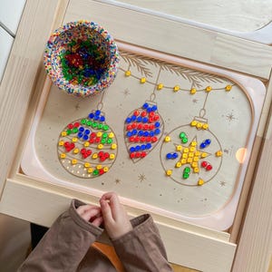 Può includere: Una tavola di attività in legno con disegni di ornamenti natalizi, riempita di pioli colorati. Una piccola ciotola contiene pioli aggiuntivi. La tavola è posta su una cornice di legno chiaro. Sono visibili le mani di un bambino.