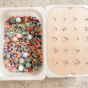 Könnte beinhalten: Ein weißes Tablett gefüllt mit bunten Pompons und ein Holzbrett mit den Zahlen 1 bis 20 darauf gedruckt.