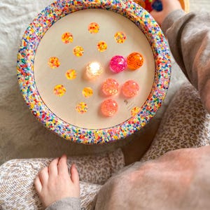 Op de afbeelding: Een houten speelgoed met een kleurrijke, met kralen bezette rand en een lichtgekleurd interieur. Het interieur bevat verschillende kleurrijke, edelsteenachtige objecten. De hand van een kind is zichtbaar, die een kleine houten kom met meer kleurrijke objecten vasthoudt.