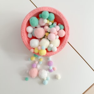 Könnte beinhalten: Eine korallenfarbene Schale gefüllt mit pastellfarbenen Pompons in Rosa, Grün, Gelb und Weiß. Einige Pompons fallen auf eine weiße Oberfläche. Das Arrangement wirkt verspielt.