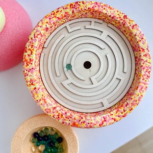 Könnte beinhalten: Ein Holz-Labyrinth-Spiel mit einer durchsichtigen Murmel im Inneren, das sich in einem farbenfrohen, gesprenkelten Behälter befindet. Eine kleine, beige Schale enthält mehrere bunte Murmeln. Der Behälter hat ein gesprenkeltes Muster aus Rosa, Gelb und Orange.