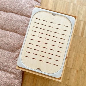 Può includere: Un vassoio di legno con una finitura in legno chiaro e un bordo in plastica bianca. Il vassoio ha un motivo a griglia di piccoli fori.