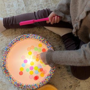 Op de afbeelding: Een rond, verlicht speelgoed met kleurrijke cirkels binnenin. Het speelgoed heeft een decoratieve rand en staat naast een kleine, gele kom. De hand van een kind houdt een roze toverstaf vast en interacteert met het speelgoed. De achtergrond is een tapijt in neutrale tinten.