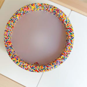 May include: A circular decorative item with a vibrant speckled design in shades of red, yellow, blue, and green. The object is placed on a white surface.
