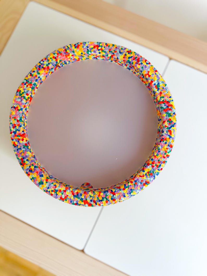 May include: A circular decorative item with a vibrant speckled design in shades of red, yellow, blue, and green. The object is placed on a white surface.