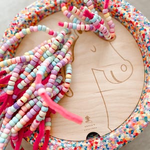 Può includere: Un giocattolo di legno a forma di unicorno con criniera di perline colorate e bordo maculato. La criniera dell'unicorno è fatta di perline color pastello infilate su scovolini rosa. Il bordo è decorato con un motivo a coriandoli.