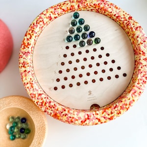 Può includere: Un gioco di biglie in legno con una base macchiata di rosso e giallo. Il gioco ha un tabellone triangolare con file numerate e buchi per far cadere le biglie. Il gioco include una ciotola di biglie e una ciotola più piccola per raccogliere le biglie.