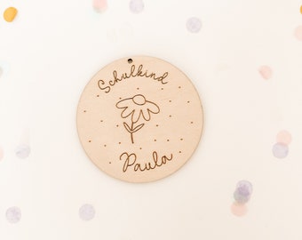 May include: A round wooden sign with the text "Schulkind" and a simple flower design. The name "Paula" is written below the flower. The sign is surrounded by confetti.