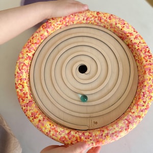 Könnte beinhalten: Ein Holz-Spiral-Labyrinth mit einem bunten, strukturierten Rand. Eine grüne Murmel befindet sich in der Mitte des Labyrinths.