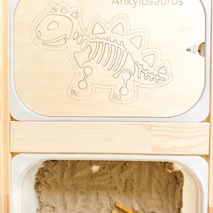 Könnte beinhalten: Ein helles Holztablett mit einem Ausschnitt eines Ankylosaurus-Skeletts. Das Tablett befindet sich über einer weißen Schale, die mit Sand und Holzpinzetten gefüllt ist. Das Wort "Ankylosaurus" ist auf das Tablett geätzt.