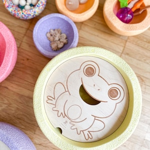Può includere: Un giocattolo di legno con un design a forma di rana e diverse ciotole colorate piene di piccoli oggetti. Le ciotole sono in tonalità di rosa, viola, arancione e blu. Il disegno della rana è inciso nel legno.