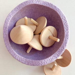 Può includere: Una ciotola viola piena di trottole di legno. Le trottole sono in legno marrone chiaro e hanno un piccolo piolo di legno al centro.