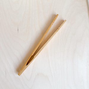 Könnte beinhalten: Hellbraune Bambuszange zum Servieren von Speisen. Die Zange hat ein einfaches, funktionales Design mit einer geschlitzten Öffnung und geriffelten Greifflächen. Die natürliche Holzmaserung ist sichtbar und verleiht ihr eine rustikale Ästhetik.