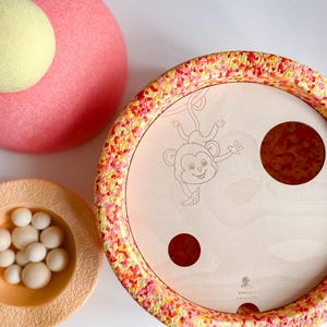 Può includere: Un giocattolo di legno con un'illustrazione di una scimmia, due cerchi rossi e un bordo maculato. Sono presenti anche un oggetto circolare rosa con un cerchio giallo sopra e una piccola ciotola con palline bianche.