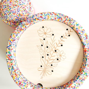 Può includere: Un vassoio di legno con un design floreale e un bordo colorato a forma di coriandoli. Il vassoio presenta un bouquet di fiori tagliato al laser e piccoli punti neri. Sullo sfondo c'è una palla abbinata ricoperta di coriandoli.