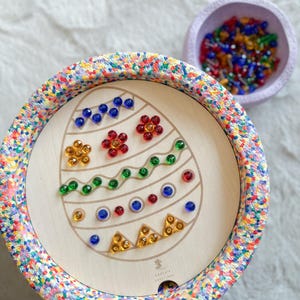 Op de afbeelding: Houten paasei-prikbord met kleurrijke pinnen. Het eierontwerp heeft blauwe, rode, groene en gele pinnen gerangschikt in bloemen- en geometrische patronen. Een kleine kom gevuld met diverse pinnen staat in de buurt.