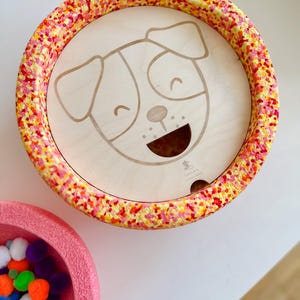 Può includere: Un cane di legno giocattolo con un viso sorridente e un buco nella bocca. Il cane è circondato da un anello macchiato di giallo, arancione e rosso. Il giocattolo è progettato per permettere ai bambini di far cadere le palline nel buco.