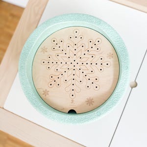 Può includere: Un giocattolo rotondo in legno verde chiaro con un design a fiocco di neve. Il centro è in legno chiaro con un motivo a fiocco di neve e piccoli fori. Il bordo esterno è verde chiaro strutturato. Il giocattolo è su una superficie bianca.