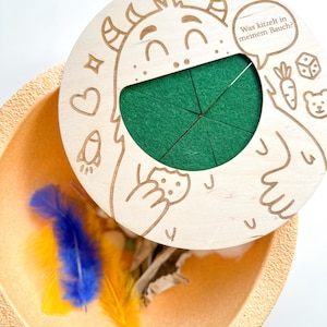 K&ouml;nnte beinhalten: Ein Holzspielzeug mit einem Monsterdesign, mit einem gr&uuml;nen Filzeinsatz und einem Deckel mit dem Text "Was kitzelt in meinem Bauch?". Das Spielzeug ist ge&ouml;ffnet und zeigt bunte Federn und andere kleine Gegenst&auml;nde in einer beigen Schale.