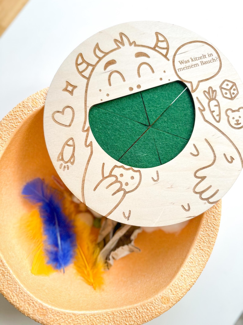 K&ouml;nnte beinhalten: Ein Holzspielzeug mit einem Monsterdesign, mit einem gr&uuml;nen Filzeinsatz und einem Deckel mit dem Text "Was kitzelt in meinem Bauch?". Das Spielzeug ist ge&ouml;ffnet und zeigt bunte Federn und andere kleine Gegenst&auml;nde in einer beigen Schale.