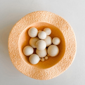 Peut inclure: Un bol de couleur pêche avec une surface texturée rempli de boules en bois de différentes tailles. Les boules sont disposées de manière aléatoire et sont toutes de la même couleur.