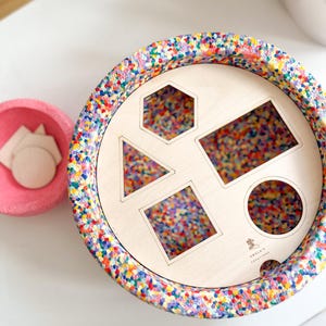 Può includere: Un giocattolo di smistamento di forme in legno con un bordo maculato colorato e un centro in legno chiaro. Il centro ha ritagli di un esagono, un triangolo, un rettangolo, un quadrato e un cerchio. Una ciotola rosa con forme di legno è accanto allo smistatore.