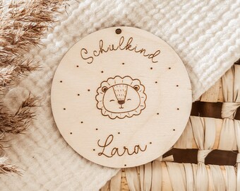 May include: A round wooden sign with the text "Schulkind" and "Lara" in cursive. A cartoon lion with a happy face is in the center of the sign.