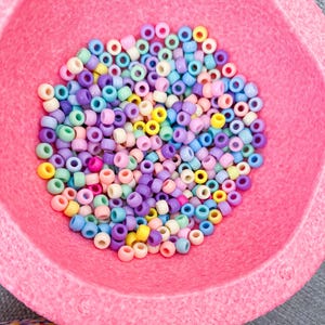 Könnte beinhalten: Eine Sammlung bunter Ponyperlen in Rosa, Lila, Gelb, Blau und Grün, die eine rosa Filzschale füllen. Diese kleinen, runden Perlen eignen sich ideal zum Basteln und für die Schmuckherstellung.
