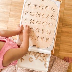 Puede incluir: Un rompecabezas de madera del alfabeto con una bandeja blanca. El rompecabezas tiene las letras del alfabeto recortadas y la bandeja contiene las piezas de las letras. Un niño está jugando con el rompecabezas.
