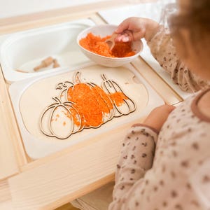 Può includere: Un puzzle di zucca in legno riempito con materiale arancione. Un bambino usa un cucchiaio di legno per riempire il puzzle. Il puzzle è su un tavolo di legno con un contenitore bianco.