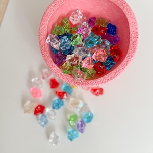 Könnte beinhalten: Eine rosa, strukturierte Schale gefüllt mit farbenfrohen, facettierten Acryl-Edelsteinen. Die Edelsteine sind in verschiedenen Farben, darunter Rot, Blau, Grün und Lila. Einige Edelsteine sind außerhalb der Schale auf einer weißen Oberfläche verstreut.