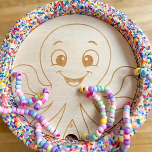Può includere: Giocattolo in legno con un design a polpo sorridente. Il bordo è ricoperto di coriandoli colorati. Diversi tentacoli di perline dai colori pastello sono disposti attorno al polpo.