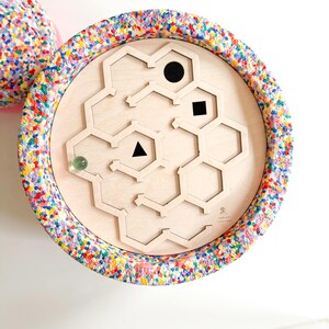 Può includere: Giocattolo labirinto in legno con finitura in legno chiaro, con un design geometrico con un cerchio, un quadrato e un triangolo neri. Il bordo esterno è rivestito in un tessuto colorato e maculato. Un biglia trasparente è all'interno del labirinto.