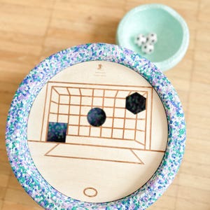 Può includere: Un gioco di calcio in legno con un bordo maculato colorato. Il tabellone presenta una porta da calcio e forme geometriche. Una piccola ciotola verde chiaro contiene mini palloni da calcio.