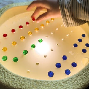 Può includere: Un tavolo luminoso sensoriale rotondo, color crema, con gemme sfaccettate colorate. Le gemme sono disposte in file di rosso, giallo, verde e blu. Una mano si allunga per prendere una gemma rossa.