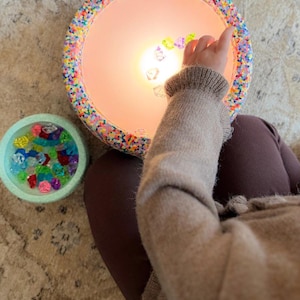 May include: A circular lamp with a colourful beaded edge, illuminated from within. A small, light blue bowl filled with colourful gems is next to the lamp. A person's arm and leg are visible.