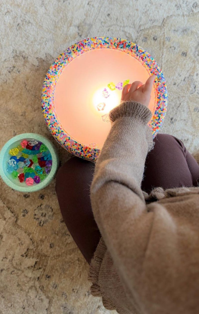 May include: A circular lamp with a colourful beaded edge, illuminated from within. A small, light blue bowl filled with colourful gems is next to the lamp. A person's arm and leg are visible.
