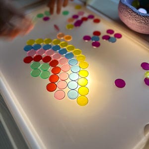 Könnte beinhalten: Ein weißer Leuchttisch mit einer Vielzahl farbiger, durchscheinender Scheiben. Die Scheiben sind in Mustern angeordnet, einige sind auf dem Tisch verstreut. Die Farben umfassen Rot, Rosa, Grün, Blau, Gelb und Orange. Der Tisch hat abgerundete Kanten.