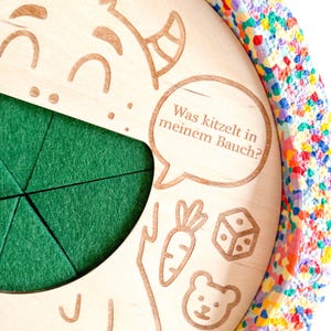 K&ouml;nnte beinhalten: Kinderspielzeug aus Holz mit einem gr&uuml;nen Filzeinsatz und einem bunten, konfettiartigen Rand. Das Spielzeug zeigt ge&auml;tzte Illustrationen einer Zeichentrickfigur, einer Karotte, eines W&uuml;rfels und eines B&auml;ren. Der Text in einer Sprechblase lautet: "Was kitzelt in meinem Bauch?"