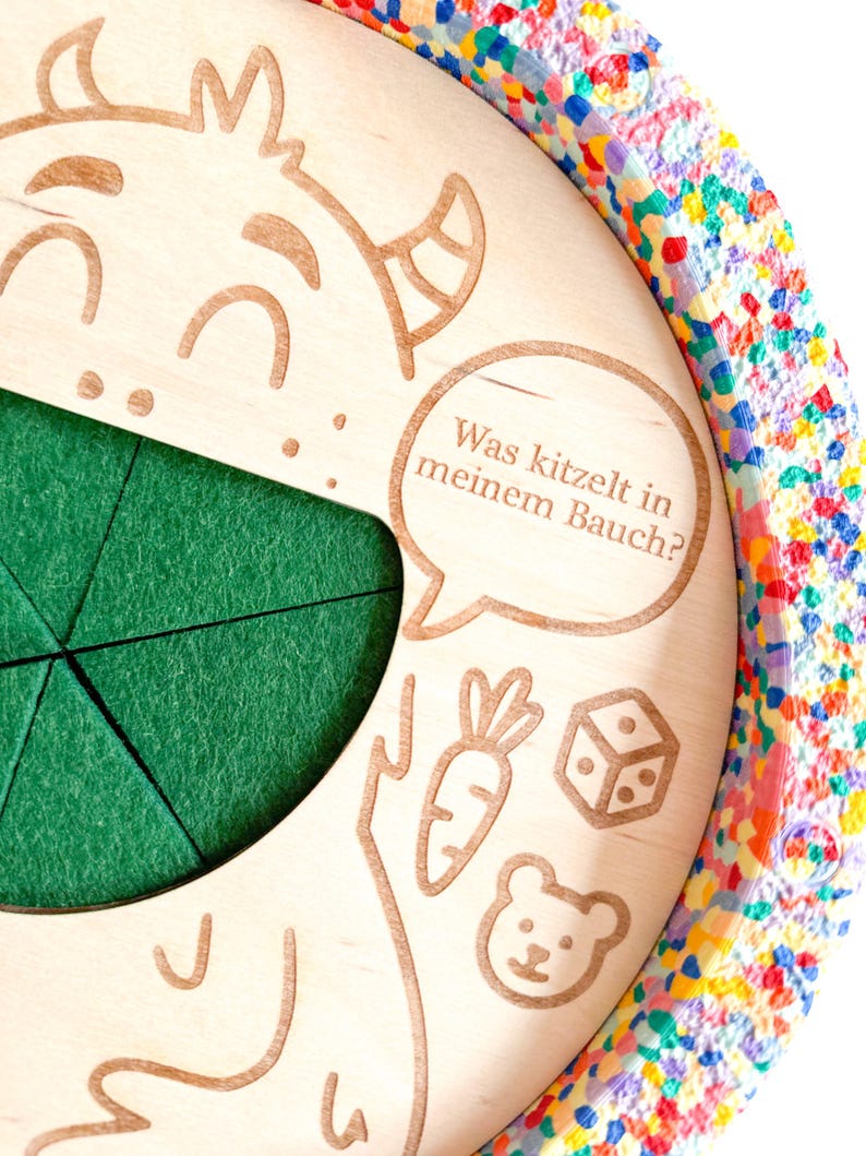 K&ouml;nnte beinhalten: Kinderspielzeug aus Holz mit einem gr&uuml;nen Filzeinsatz und einem bunten, konfettiartigen Rand. Das Spielzeug zeigt ge&auml;tzte Illustrationen einer Zeichentrickfigur, einer Karotte, eines W&uuml;rfels und eines B&auml;ren. Der Text in einer Sprechblase lautet: "Was kitzelt in meinem Bauch?"