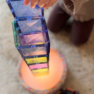 May include: A child's hand interacting with a tower of colourful, translucent magnetic tiles. The tower is composed of various colours, including blue, green, pink, and yellow. The base of the tower rests on a circular, illuminated surface.