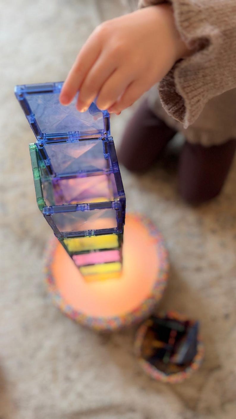 May include: A child's hand interacting with a tower of colourful, translucent magnetic tiles. The tower is composed of various colours, including blue, green, pink, and yellow. The base of the tower rests on a circular, illuminated surface.